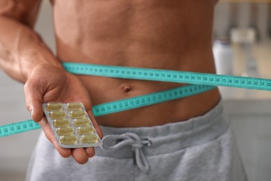 Photo of Athletic man holding pills and measuring his waist with tape indoors, closeup. Weight loss