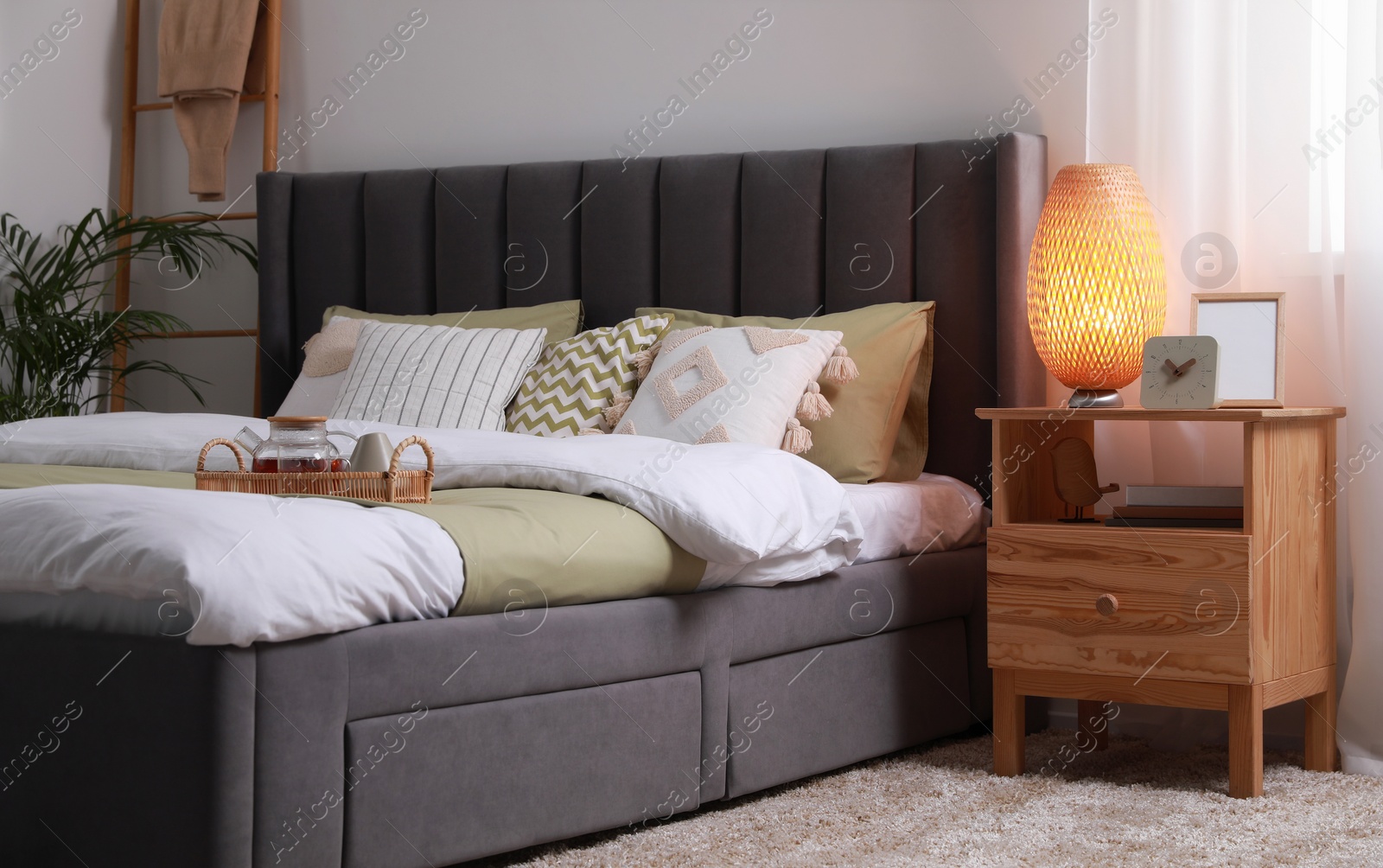 Photo of Wicker tray with teapot on comfortable bed, lamp and different decor on wooden bedside table in room. Stylish interior