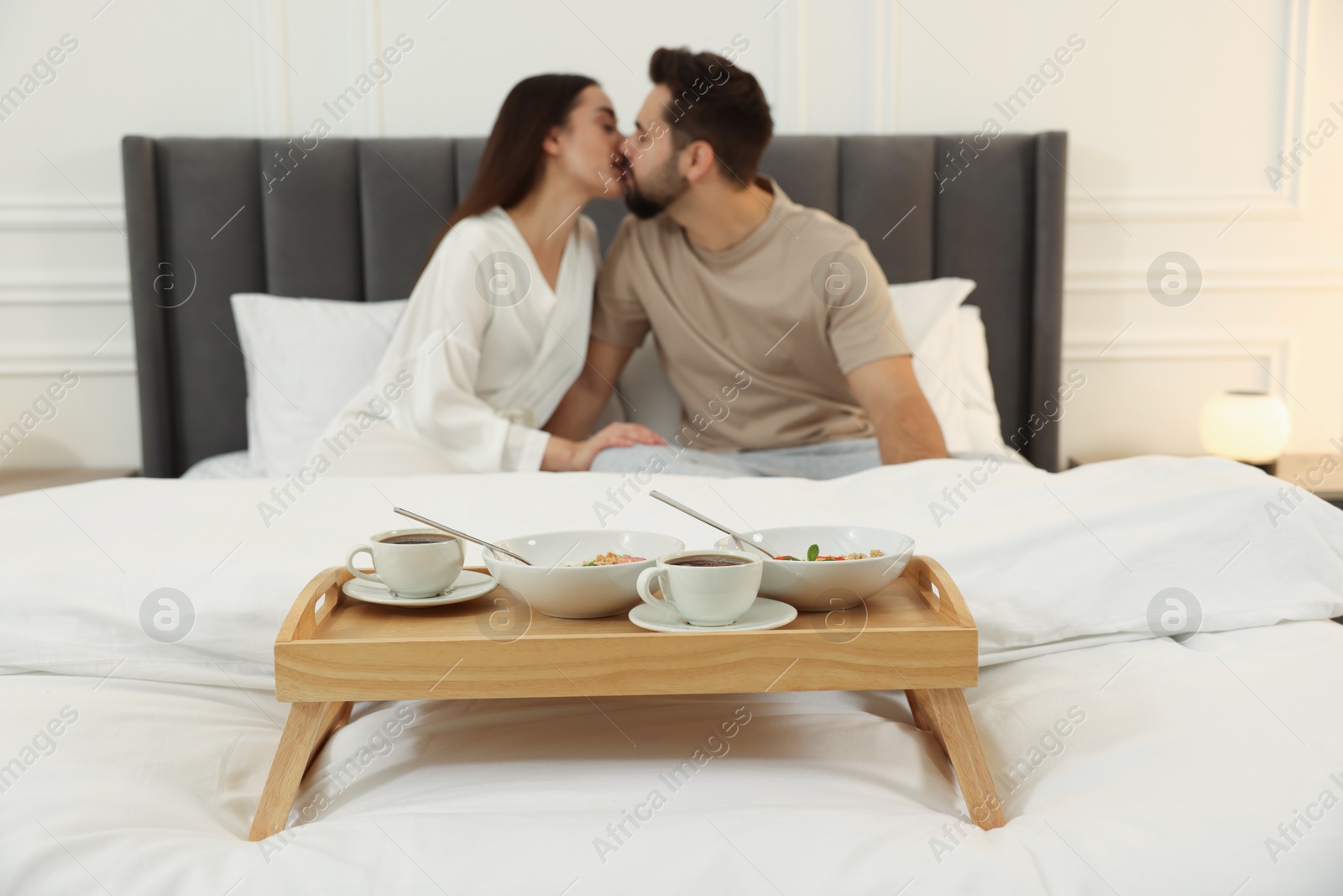 Photo of Happy couple kissing on bed at home, focus on wooden tray with breakfast