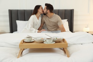 Happy couple kissing on bed at home, focus on wooden tray with breakfast