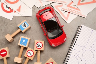 Photo of Many different road signs, notebook with sketch of roundabout and toy car on grey table, flat lay. Driving school