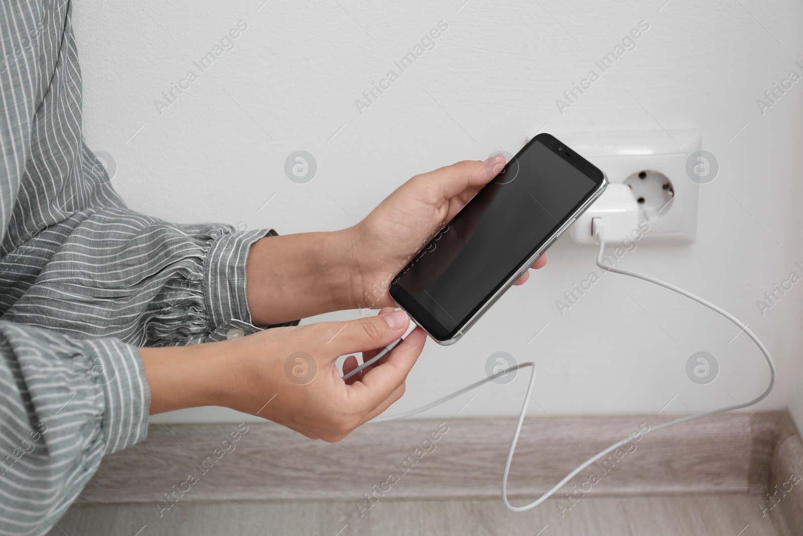 Photo of Woman connecting charger cable to mobile phone near white brick wall indoors, closeup