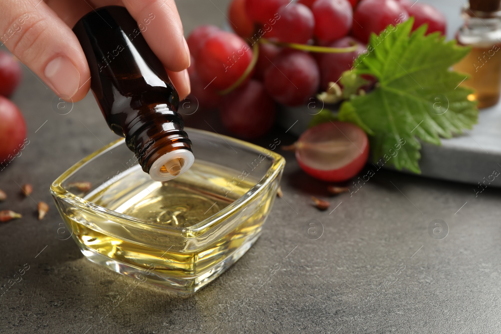 Photo of Woman dripping natural grape seed oil into bowl at grey table, closeup. Organic cosmetic