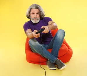 Emotional mature man playing video games with controller on color background