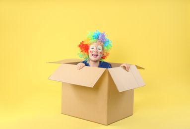 Little boy in clown wig sitting inside of cardboard box on yellow background. April fool's day