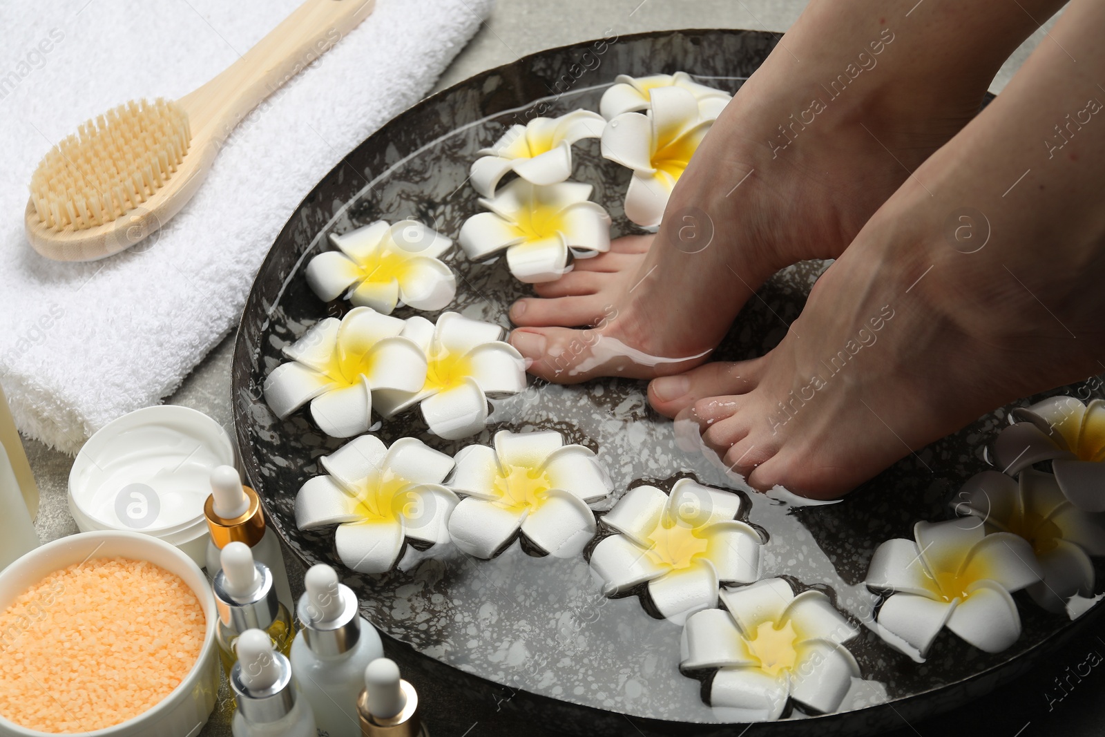 Photo of Woman soaking her feet in bowl with water and flowers on floor, closeup. Spa treatment