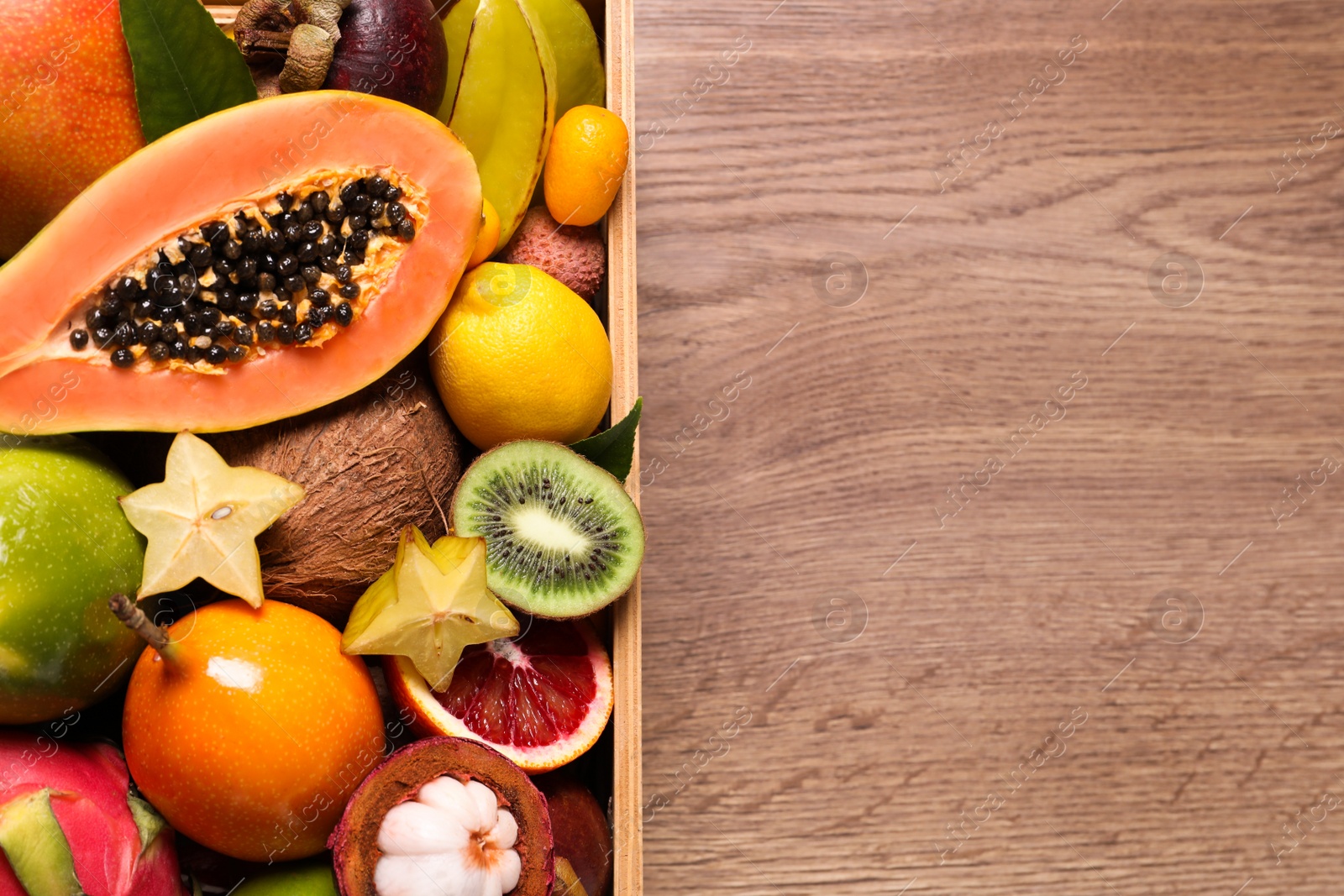 Photo of Different tropical fruits in box on wooden table, top view. Space for text