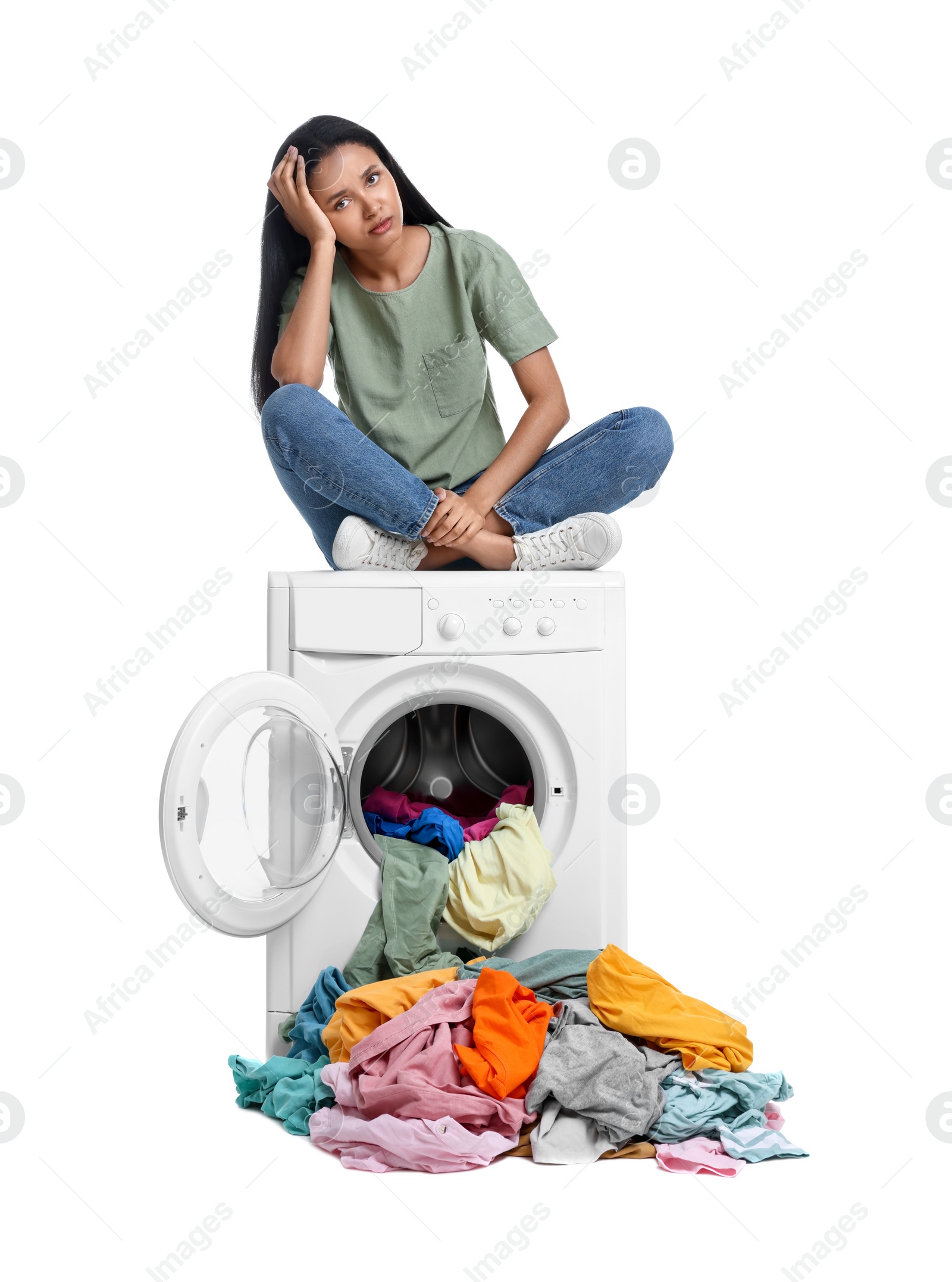 Photo of Confused woman sitting on washing machine with pile of laundry against white background