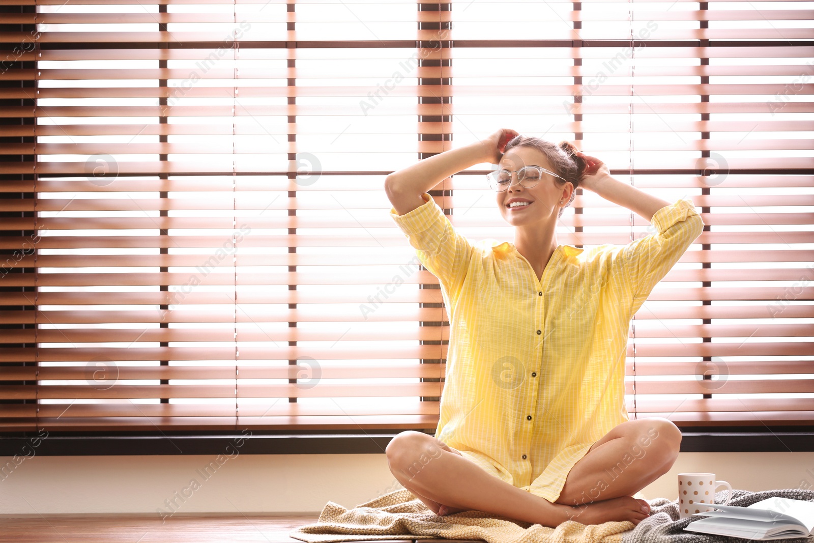 Photo of Young woman reading book near window with blinds at home. Space for text