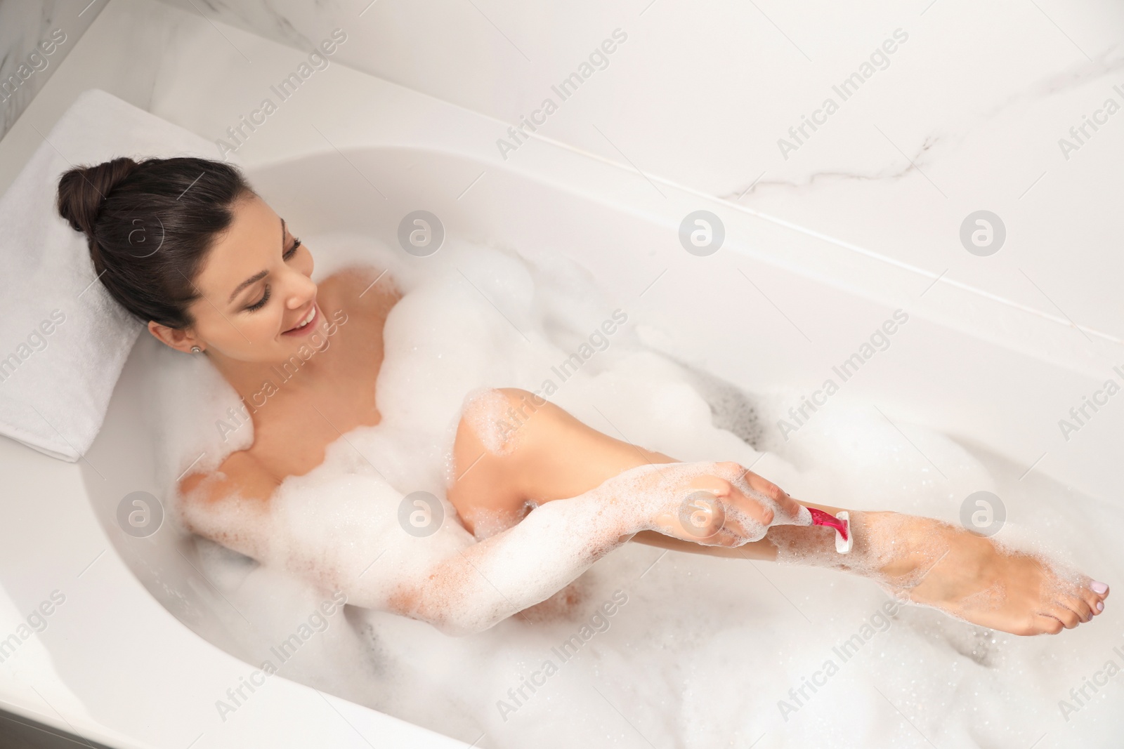 Photo of Young woman shaving legs in bubble bath, above view