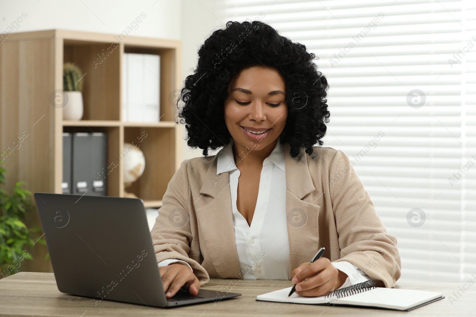 Photo of Happy young woman writing notes while using laptop at wooden desk indoors