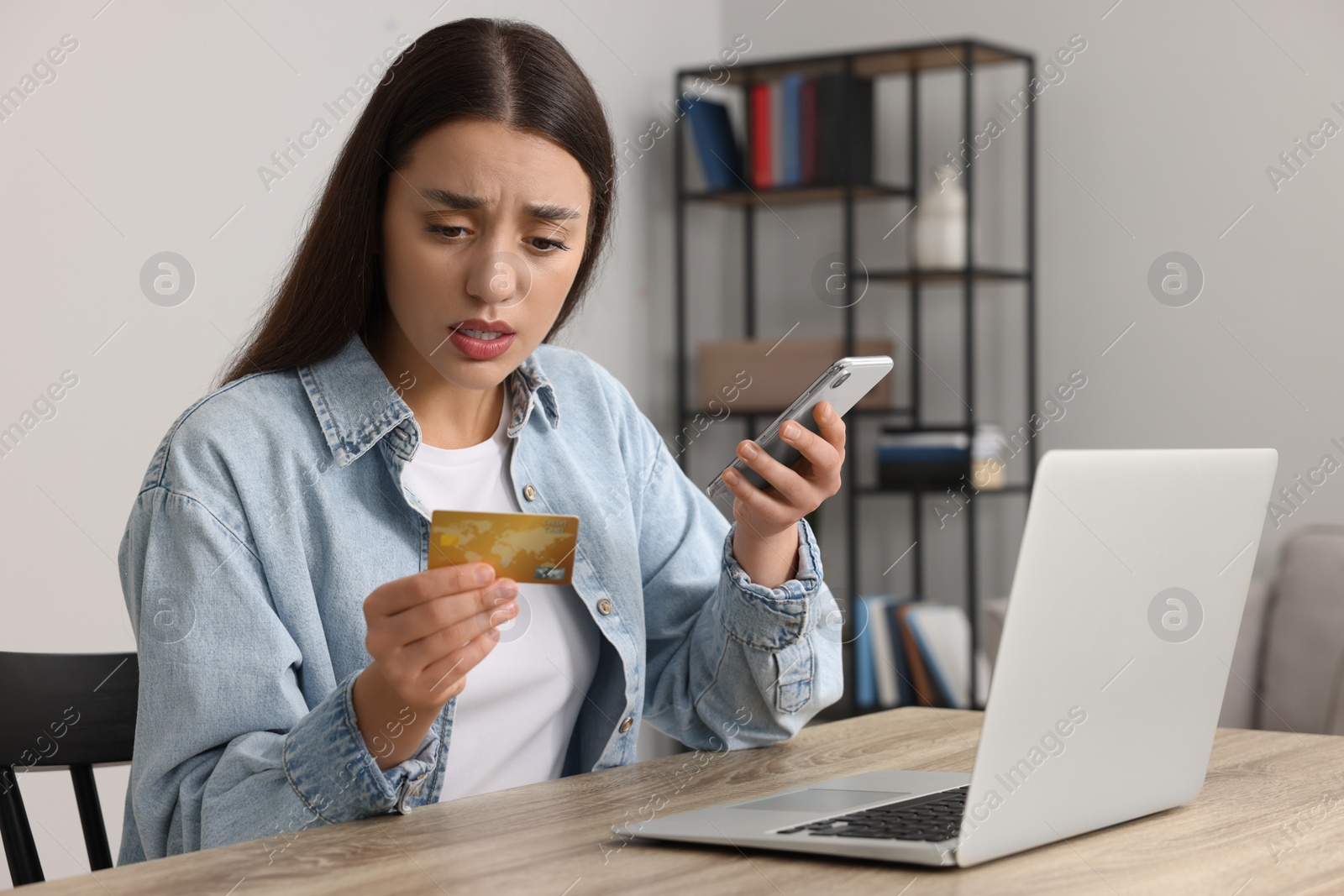 Photo of Confused woman with credit card, smartphone and laptop at table indoors. Be careful - fraud