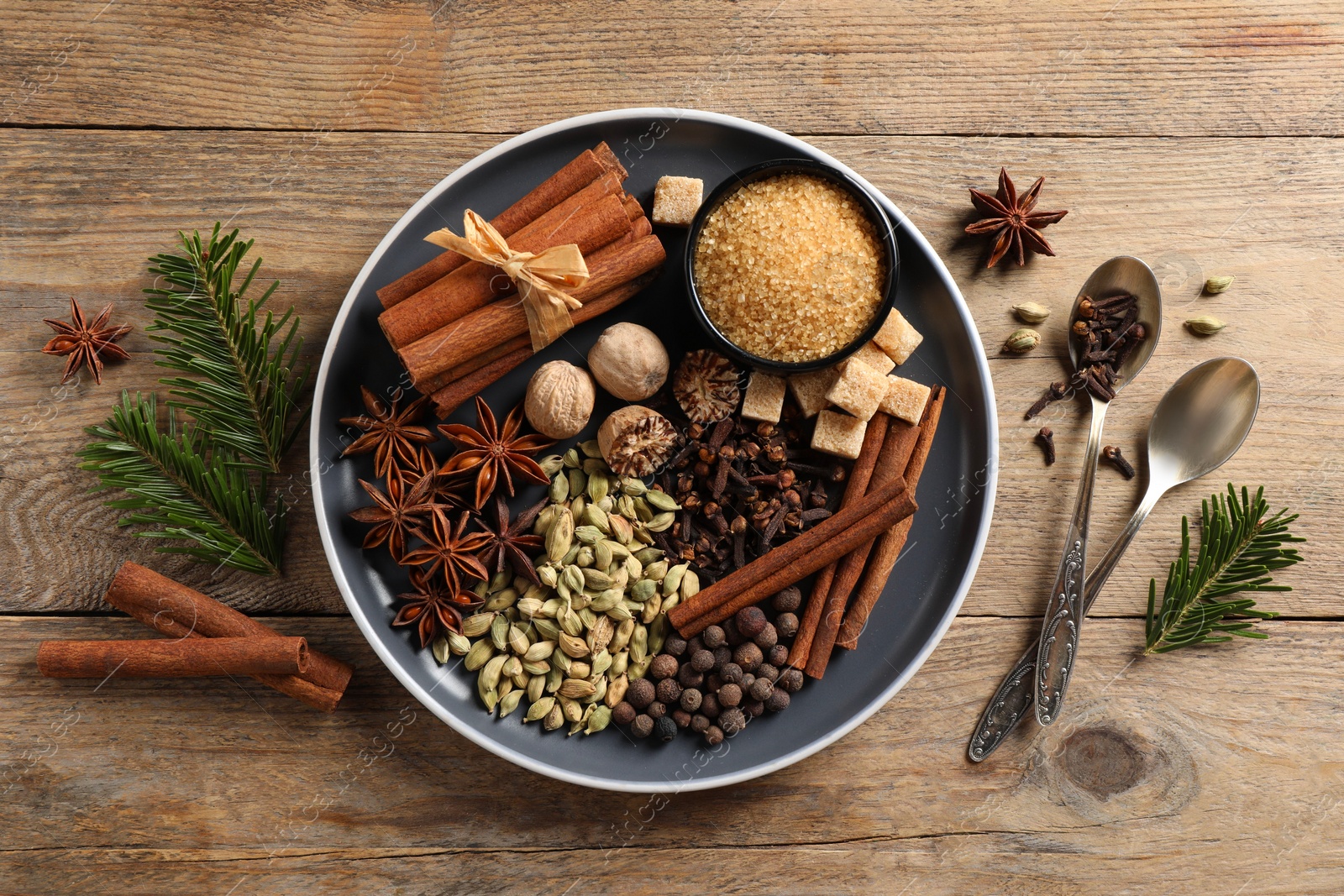 Photo of Plate with different aromatic spices, spoons and fir branches on wooden table, flat lay