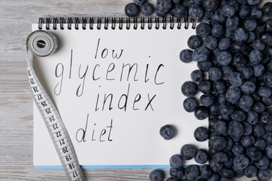 Photo of Notebook with words Low Glycemic Index Diet, measuring tape and blueberries on light wooden table, flat lay