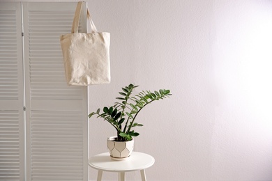 Photo of Eco bag on folding screen near table with houseplant against white wall. Space for design