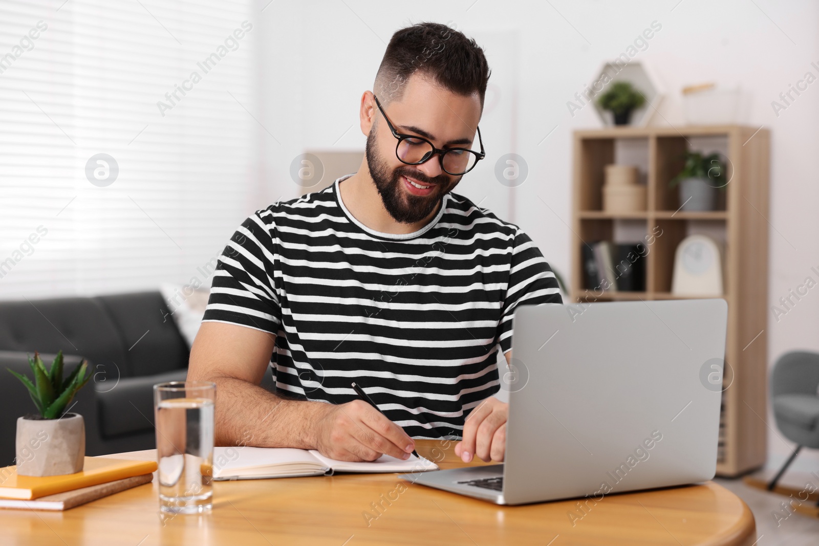 Photo of Young man writing in notebook while working on laptop at wooden table indoors