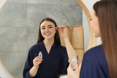 Photo of Beautiful woman in blue robe applying face serum near mirror indoors