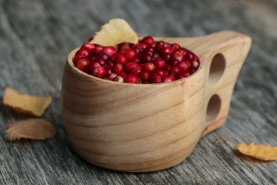Photo of Cup with tasty ripe lingonberries and leaves on wooden surface, closeup