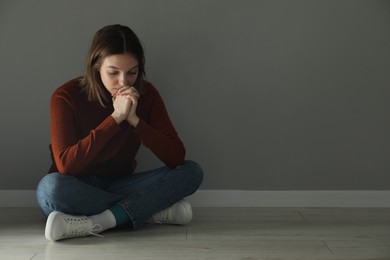 Sad young woman sitting on floor near grey wall indoors, space for text