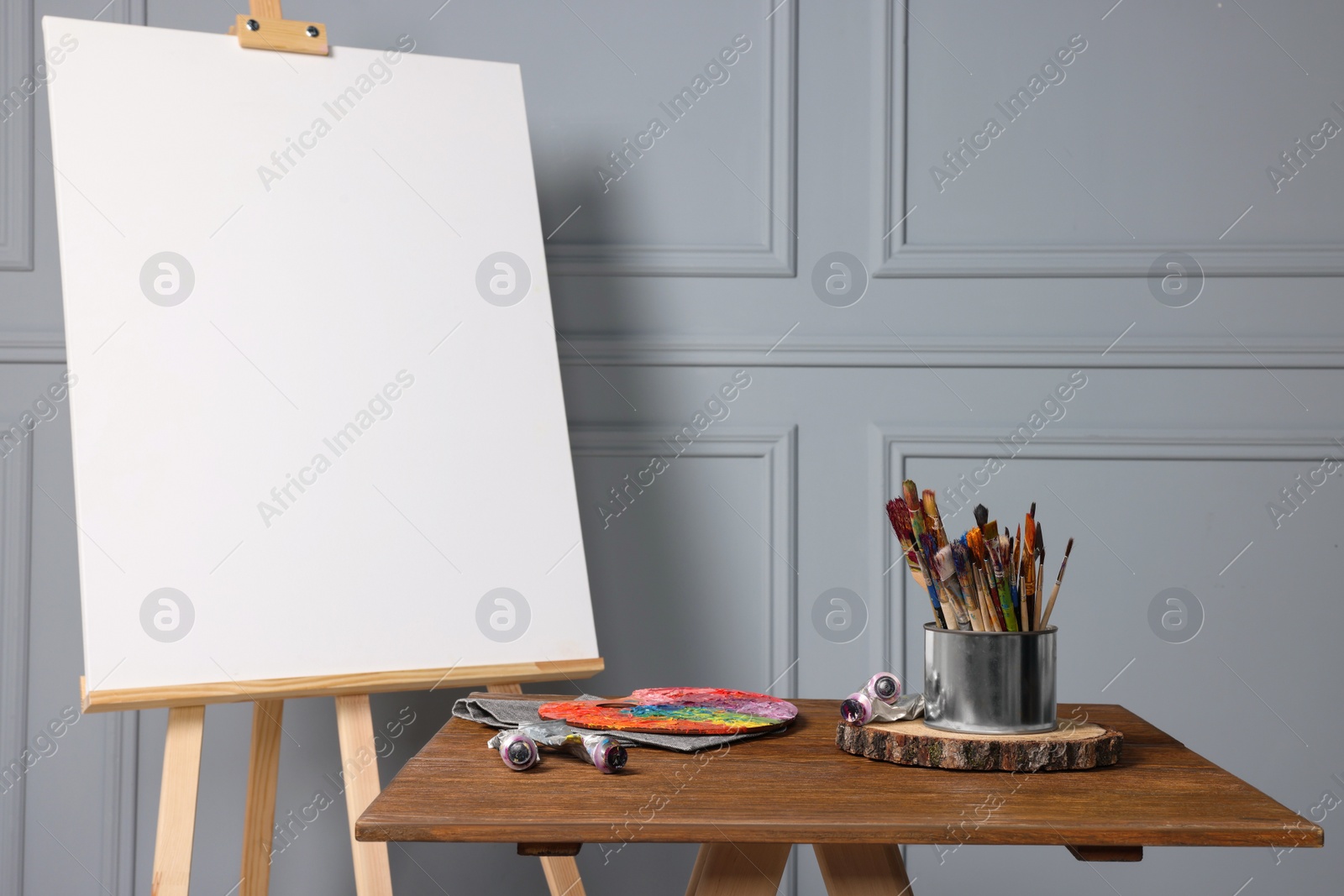 Photo of Easel with blank canvas and different art supplies on wooden table near grey wall