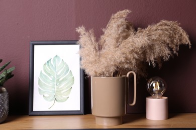 Ceramic vase with fluffy dry plants, painting and modern night lamp on wooden table near brown wall