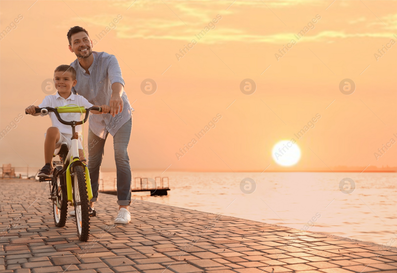 Photo of Happy father teaching his son to ride bicycle near river at sunset