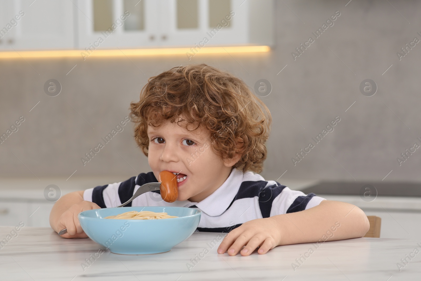 Photo of Cute little boy eating sausage and pasta at table in kitchen