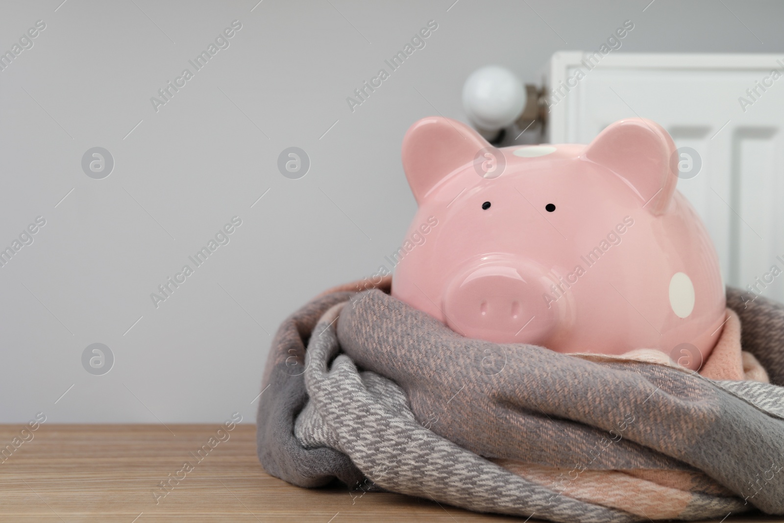 Photo of Piggy bank wrapped in scarf on wooden table near heating radiator, space for text