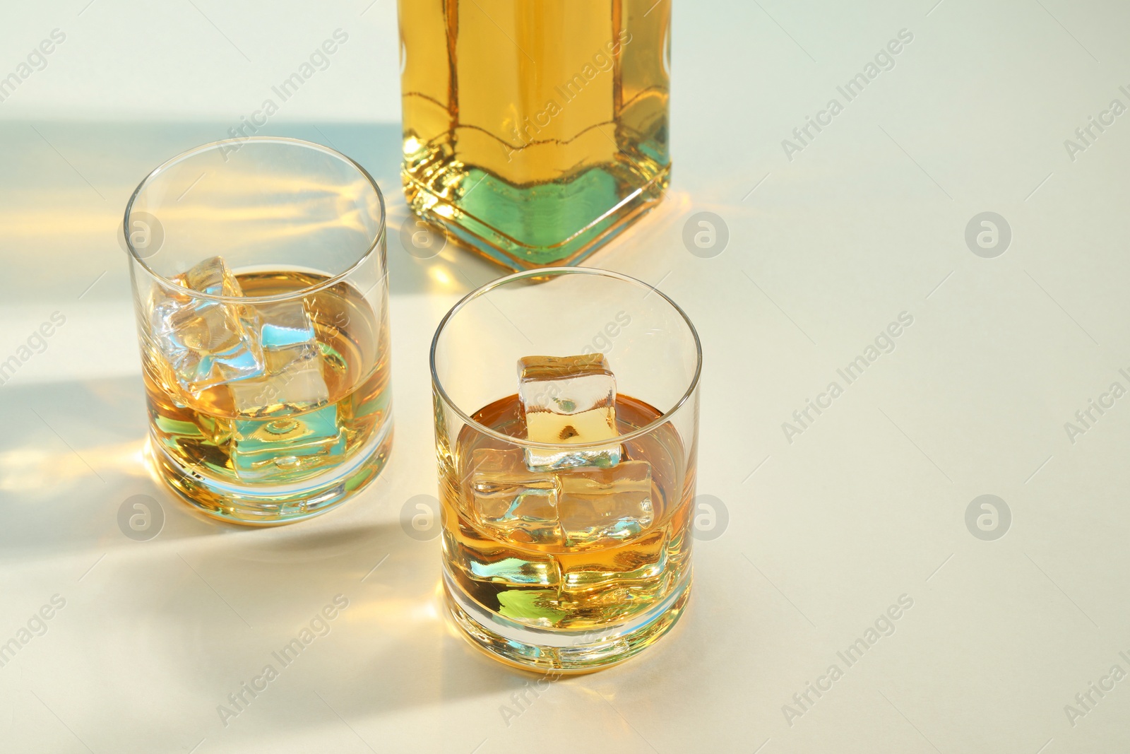 Photo of Whiskey with ice cubes in glasses and bottle on white table, space for text