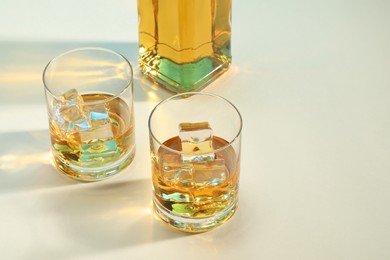 Whiskey with ice cubes in glasses and bottle on white table, space for text