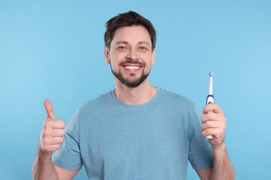 Photo of Happy man holding electric toothbrush and showing thumb up on light blue background