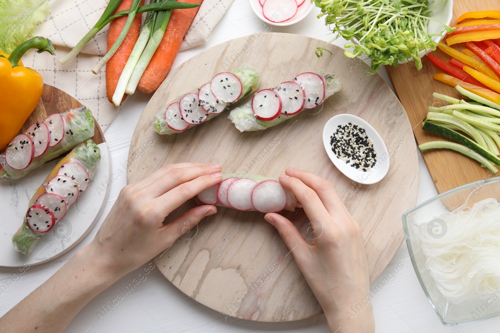 Photo of Woman wrapping spring roll at white wooden table with products, top view