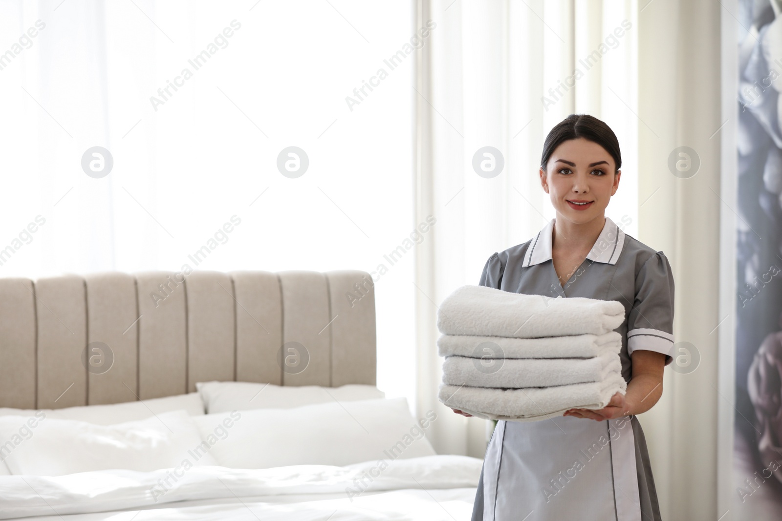 Photo of Young chambermaid holding stack of fresh towels in bedroom. Space for text