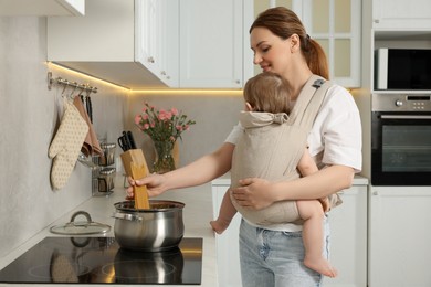 Mother holding her child in sling (baby carrier) while cooking pasta in kitchen