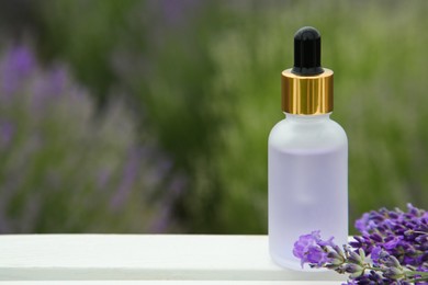 Photo of Bottle of essential oil and lavender flowers on white wooden table in field, space for text