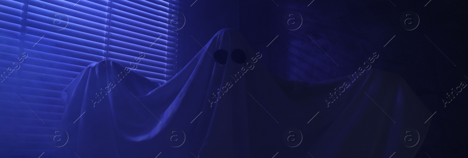 Photo of Creepy ghost. Woman covered with sheet near window in blue light