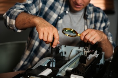 Repairman with magnifying glass and screwdriver fixing modern printer indoors, closeup