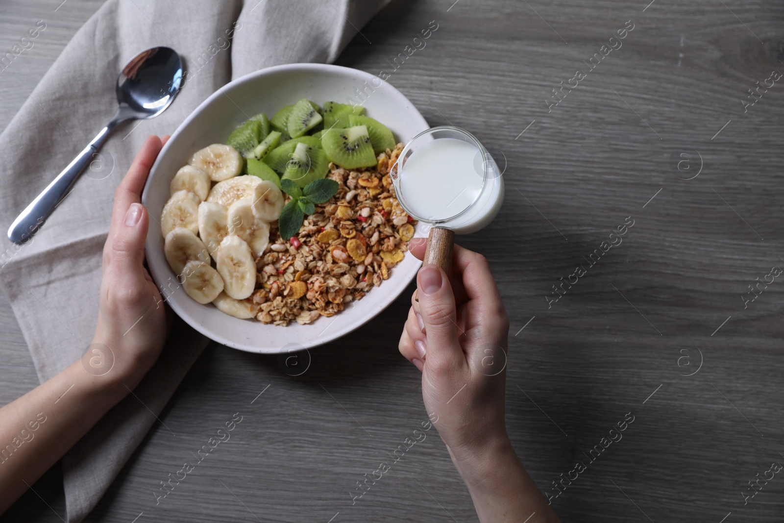 Photo of Woman pouring milk into bowl of tasty granola with banana and kiwi at grey wooden table, top view