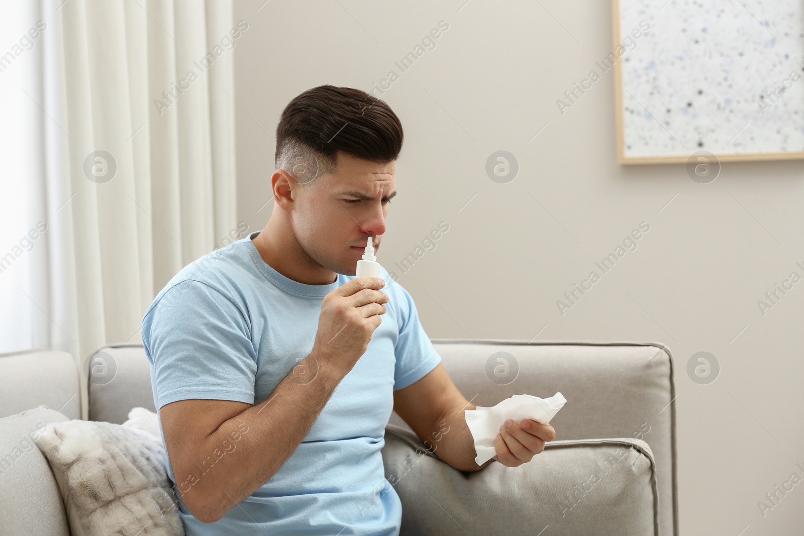 Photo of Ill man with paper tissue using nasal spray on sofa at home