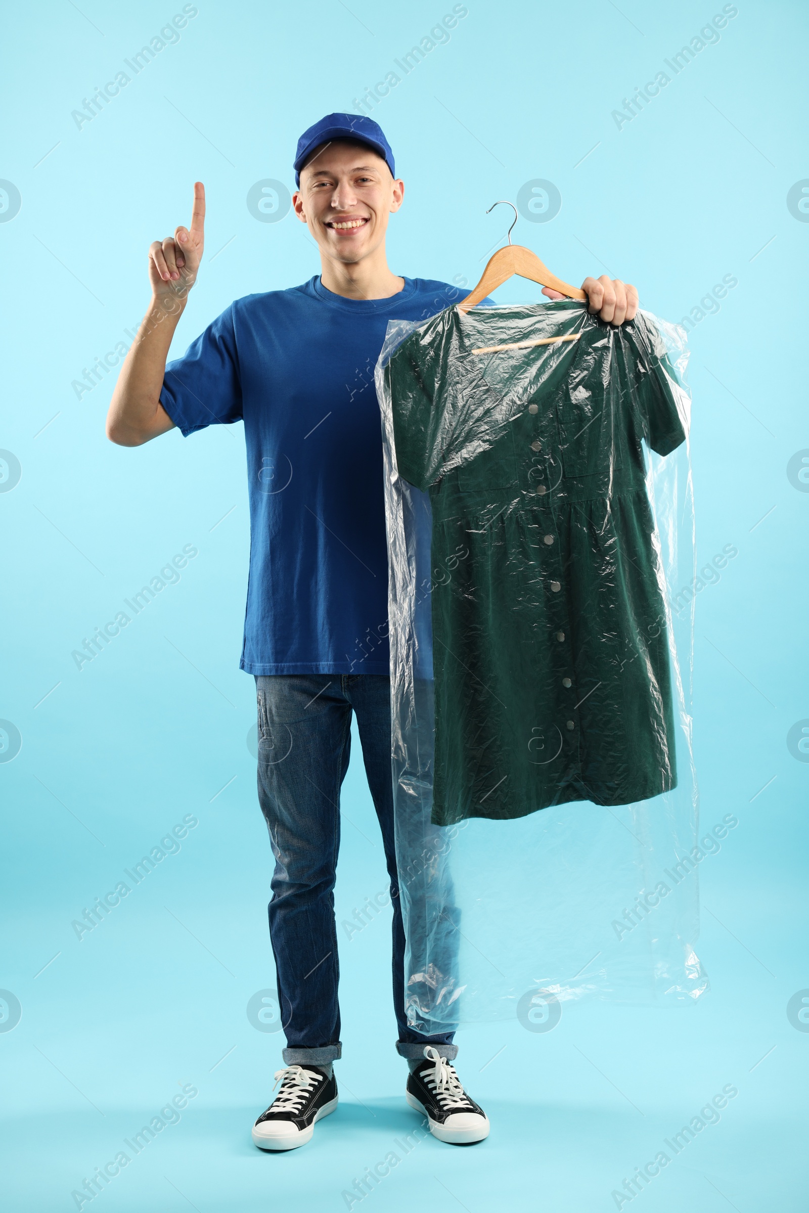 Photo of Dry-cleaning delivery. Happy courier holding dress in plastic bag and pointing at something on light blue background