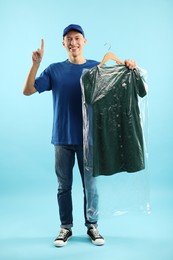 Photo of Dry-cleaning delivery. Happy courier holding dress in plastic bag and pointing at something on light blue background