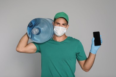 Photo of Courier in medical mask with bottle for water cooler showing mobile phone on light grey background. Delivery during coronavirus quarantine