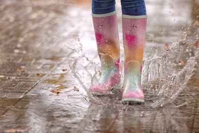 Photo of Woman wearing rubber boots splashing in puddle after rain, focus on legs. Autumn walk