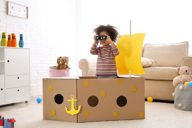Photo of Cute African American child playing with cardboard ship and binoculars at home