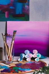 Photo of Canvas with gradient painting, brushes in glass and wooden artist's palette on table