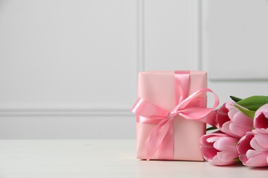 Photo of Beautiful gift box with bow and pink tulips on white table. Space for text