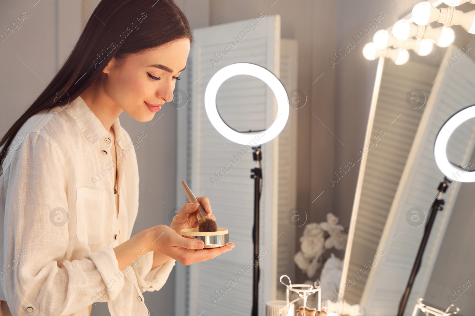 Photo of Young woman applying make up near illuminated mirror indoors