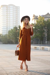 Beautiful young woman in stylish red dress and hat with handbag on city street