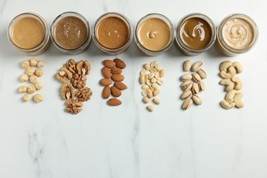 Tasty nut butters in jars and raw nuts on white marble table, flat lay. Space for text