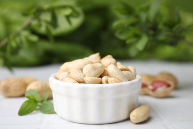 Photo of Fresh peeled peanuts in bowl on white tiled table against blurred green background, closeup